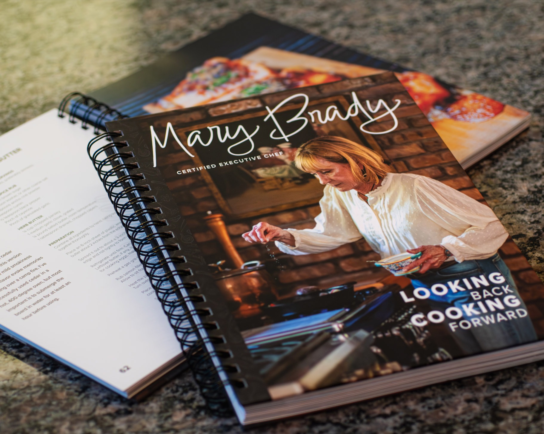 Photo of the Mary Brady Cookbook laying open on a marble table with a second cookbook showing the cover laying offset on the opened book.